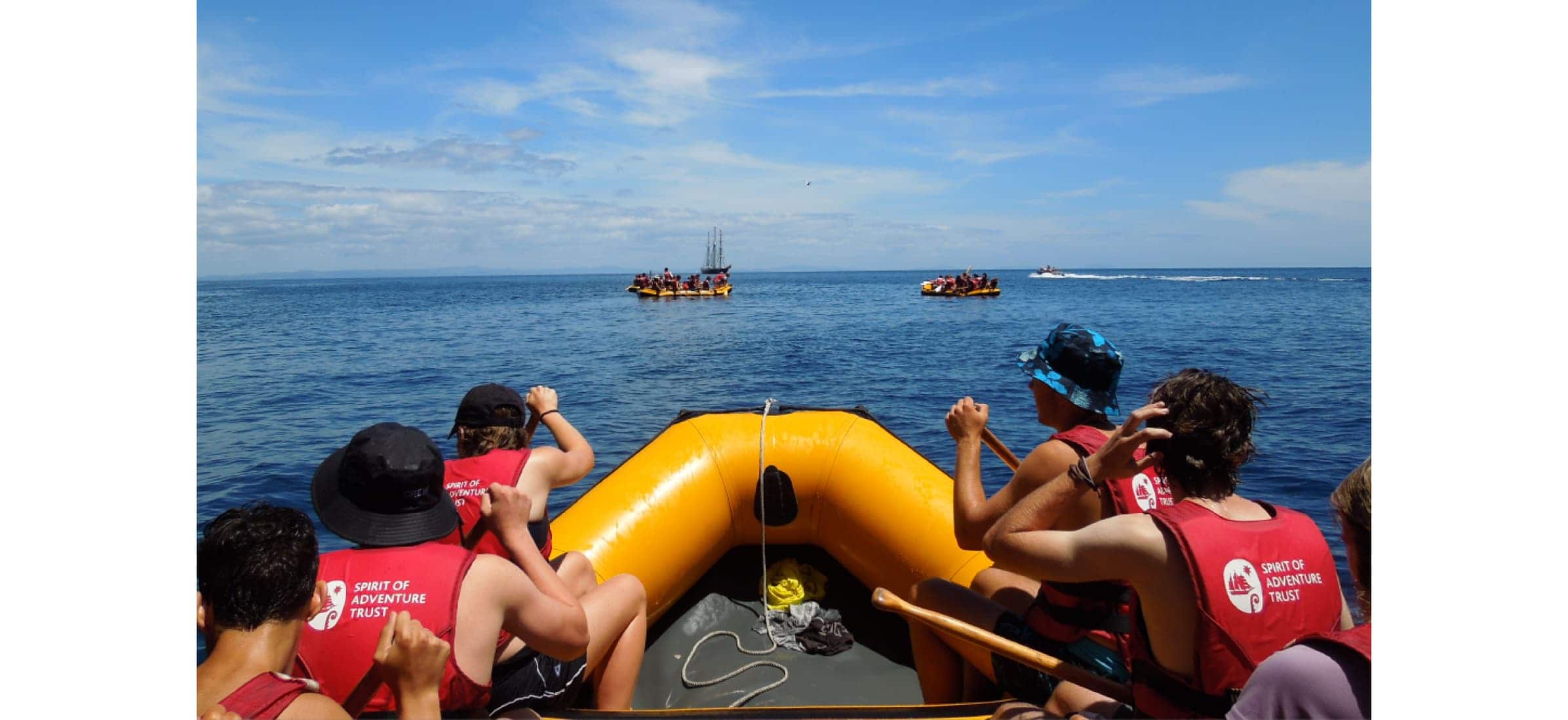 Passengers on an inflatable boat row towards two other boats, the Spirit of Adventure in the distance.